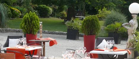An outdoor dining area with tables and chairs, surrounded by greenery and a statue.