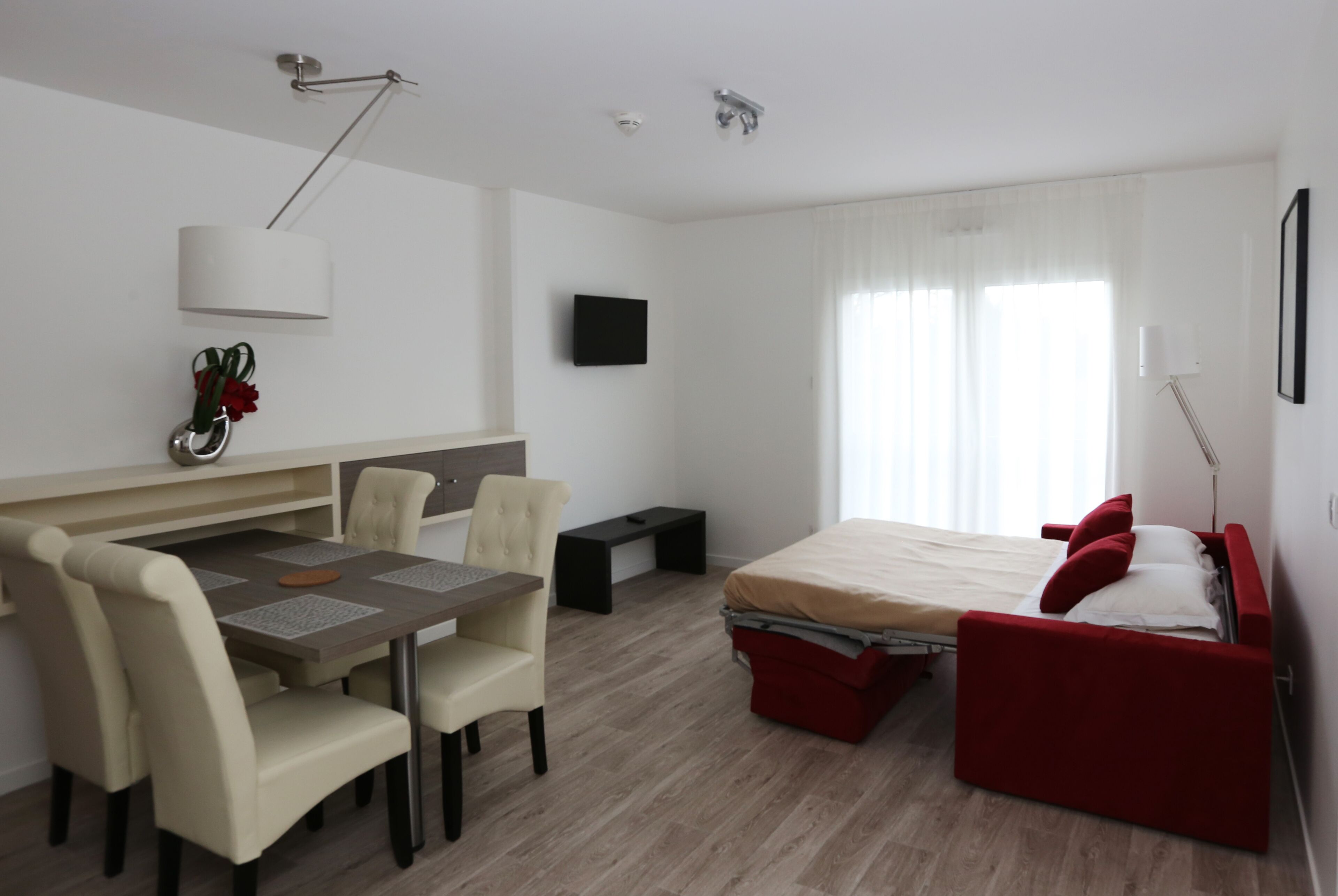 A compact hotel room with a bed, a desk with a chair, a small table, and a TV mounted on the wall.
