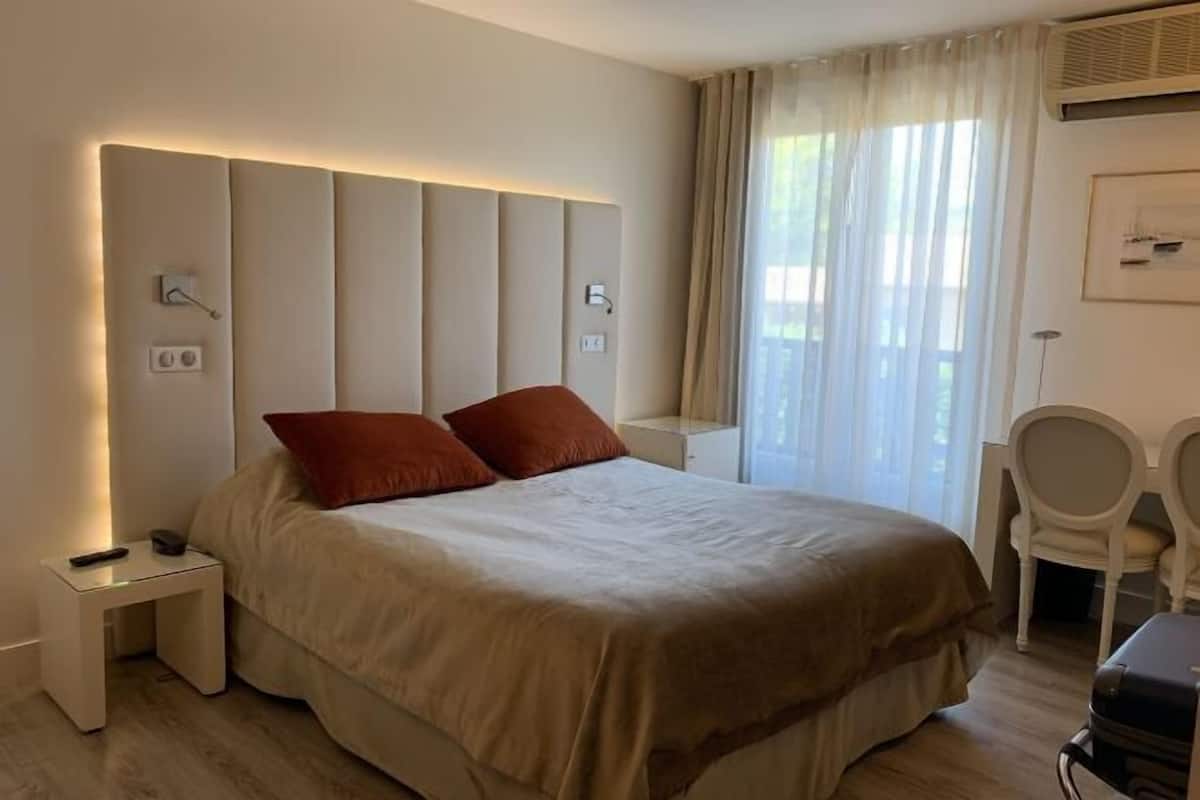 Une chambre d’hôtel moderne avec un grand lit, deux oreillers rouges, une table de chevet avec une télécommande, une chaise et un bureau avec une valise.