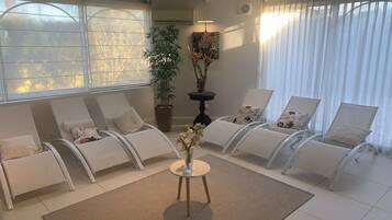 Un espace de détente moderne avec des fauteuils blancs, une table centrale agrémentée d’un vase de fleurs et une grande fenêtre ornée de rideaux légers.