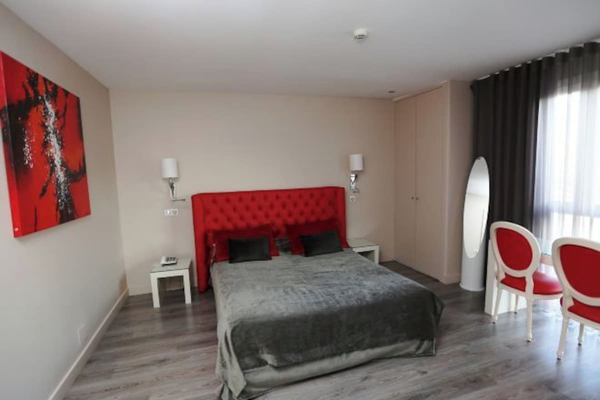 Una habitación de hotel con un cabecero tapizado en rojo, una colcha gris, dos mesitas de noche con lámparas, una silla blanca y una tabla de surf apoyada en la pared.