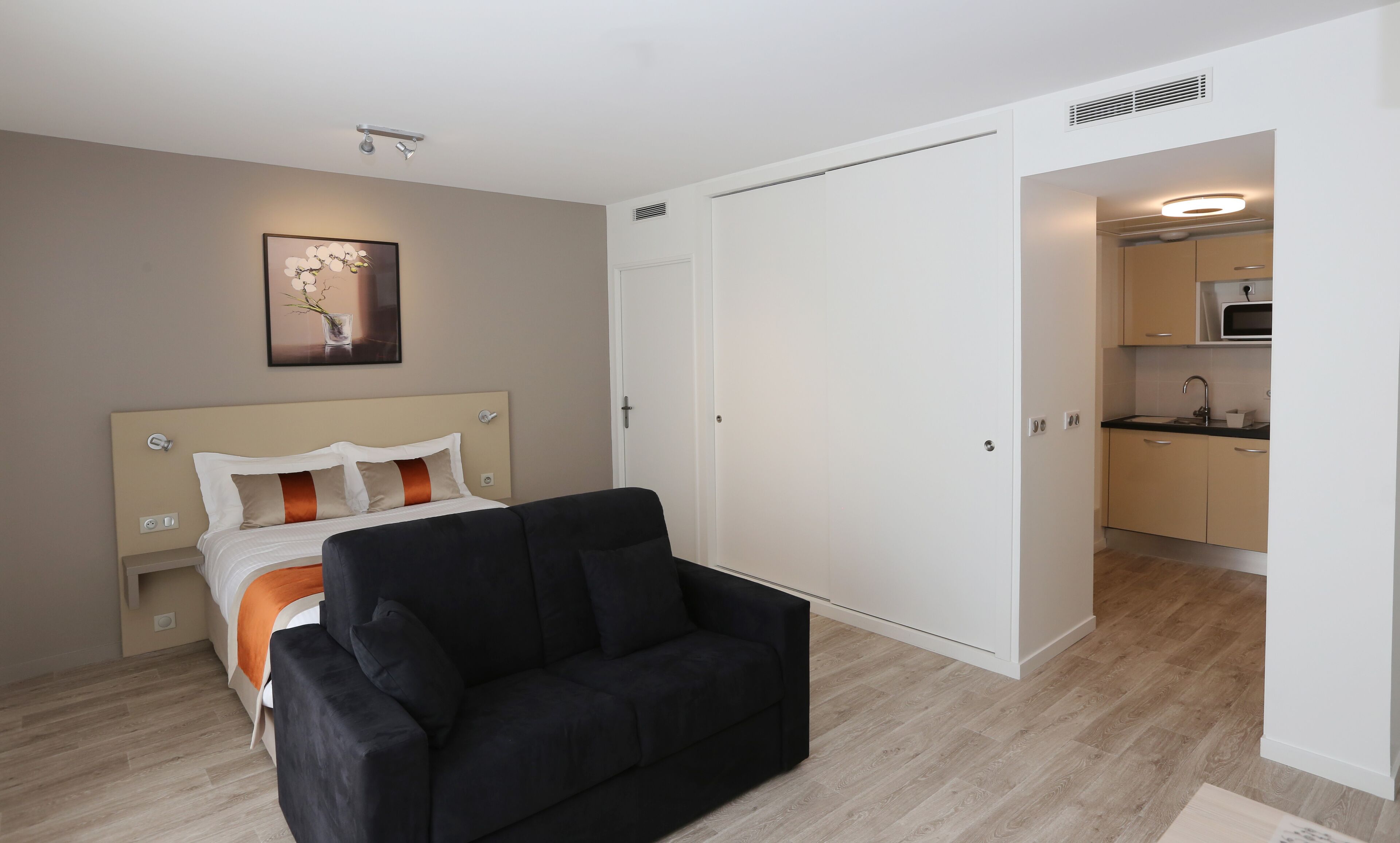 A modern hotel room with a bed, a black sofa, a painting, and a kitchenette.