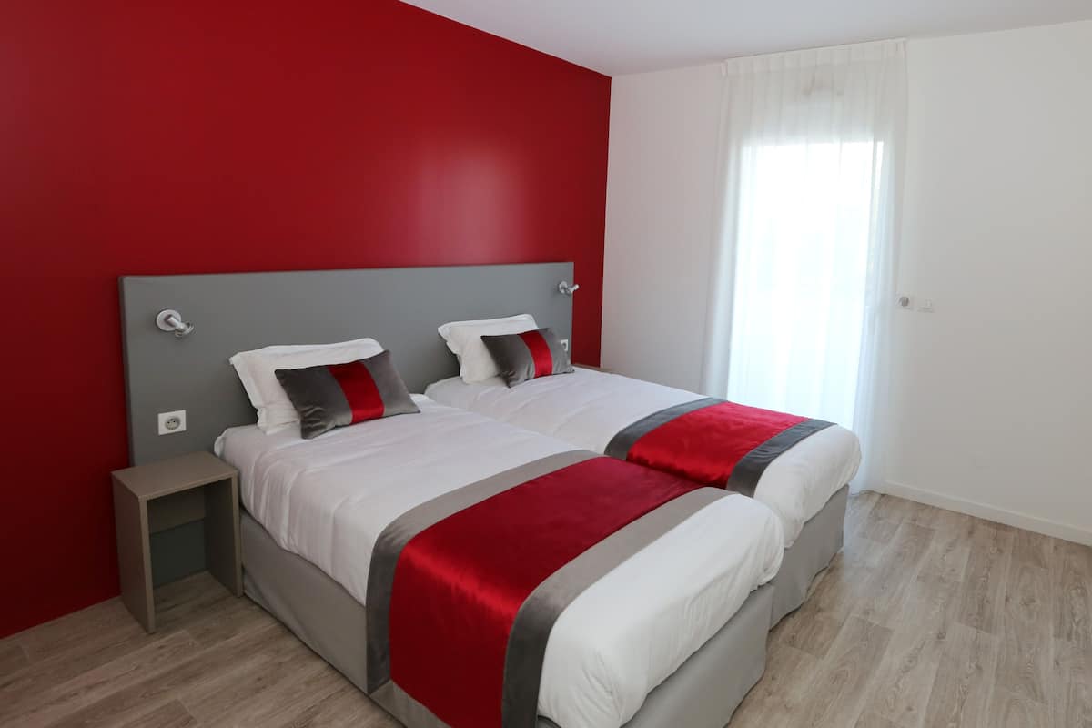Un lit double avec une tête de lit grise, une literie blanche et des touches de couleur rouge. On y trouve une table de chevet avec une lampe, une fenêtre avec des rideaux légers et un plancher en bois.