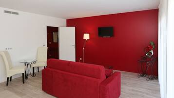 Ein Zimmer mit einem roten Sofa, einem weißen Sessel, einem kleinen Tisch, einer Stehlampe, einem an der Wand montierten Fernseher und einer Zierpflanze.