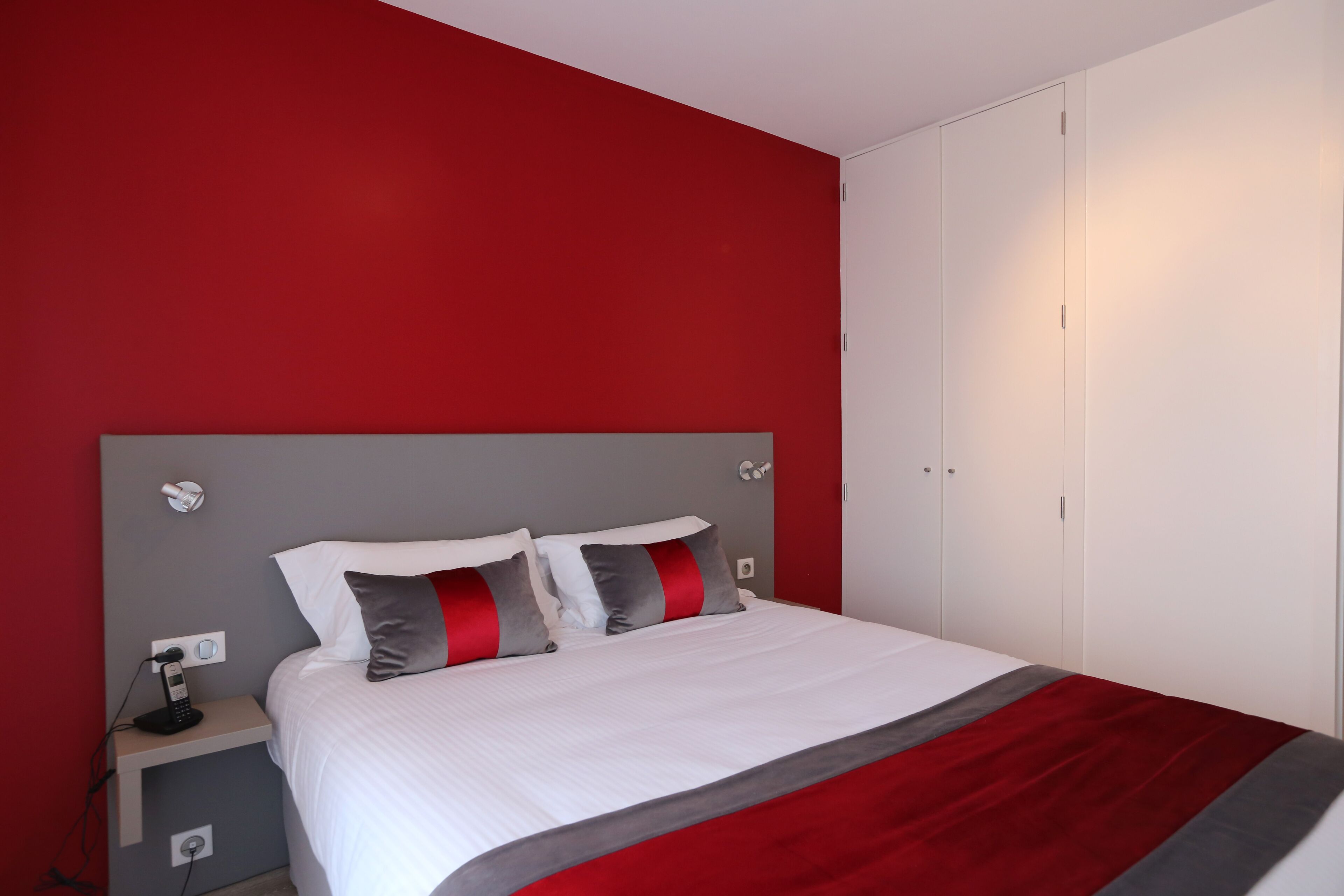 Een net opgemaakt bed met wit en grijs beddengoed, een rode accentkleur en een grijs hoofdbord. Er staat een nachtkastje met een lamp en een telefoon. De kamer heeft rode wanden en witte kasten.