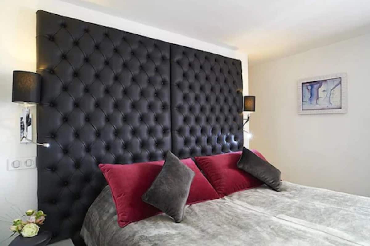 Une chambre à coucher avec une tête de lit capitonnée, des coussins rouges et gris, et une table de chevet avec une lampe.