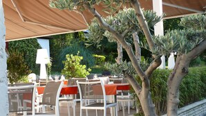 Un espacio de patio al aire libre con sillas y mesas blancas, cubierto por una sombrilla grande, rodeado de follaje y un pequeño árbol.