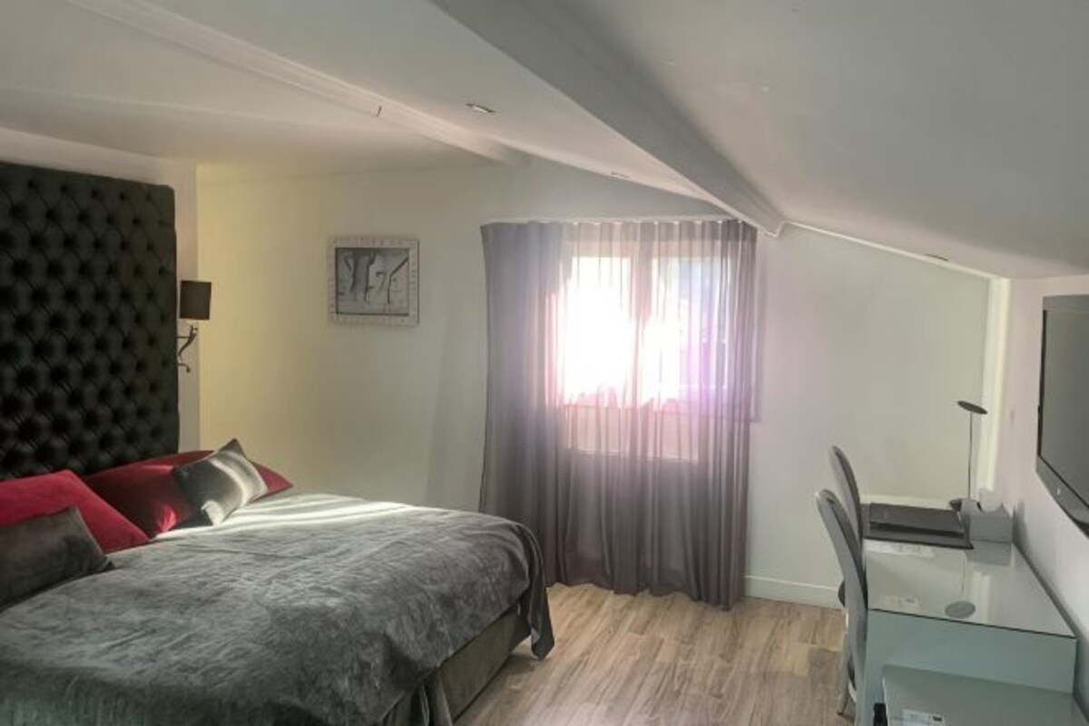 A bedroom with a bed, a desk, a chair, a TV, a window with curtains, and a clock on the wall.