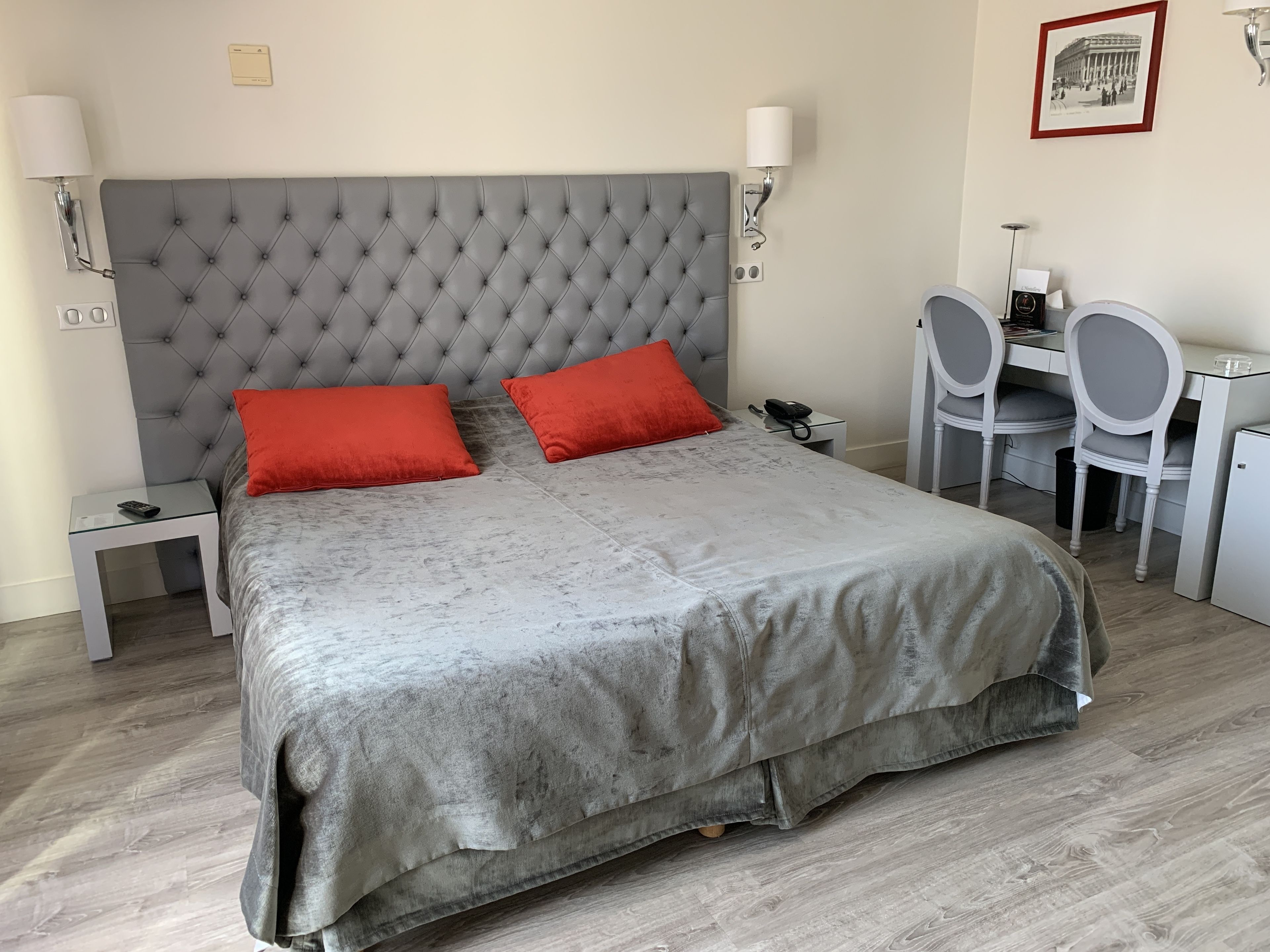 A neatly arranged bedroom with a tufted headboard, two red pillows, a bedside table with a lamp, and a desk with a chair.
