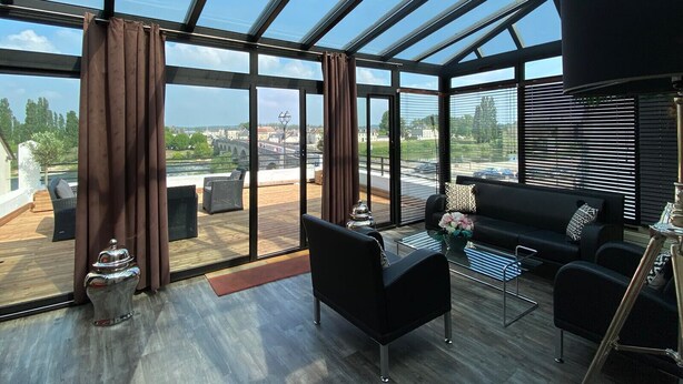 A modern interior with a glass ceiling, a black sofa, and a dining area with a view of a bridge and greenery. - Hôtel Bellevue (Amboise)