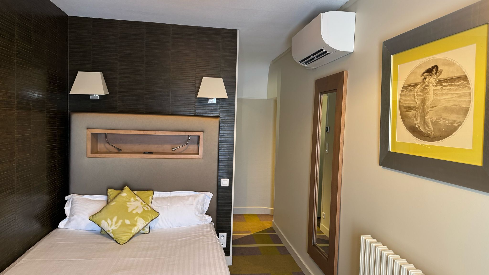 Habitación de hotel con cama, dos lámparas de noche, una obra de arte enmarcada, un equipo de aire acondicionado y un radiador.