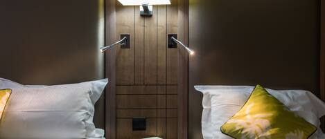Habitación de hotel con dos camas, cada una con una almohada y una mesita de noche. Hay una lámpara en la pared entre las camas.