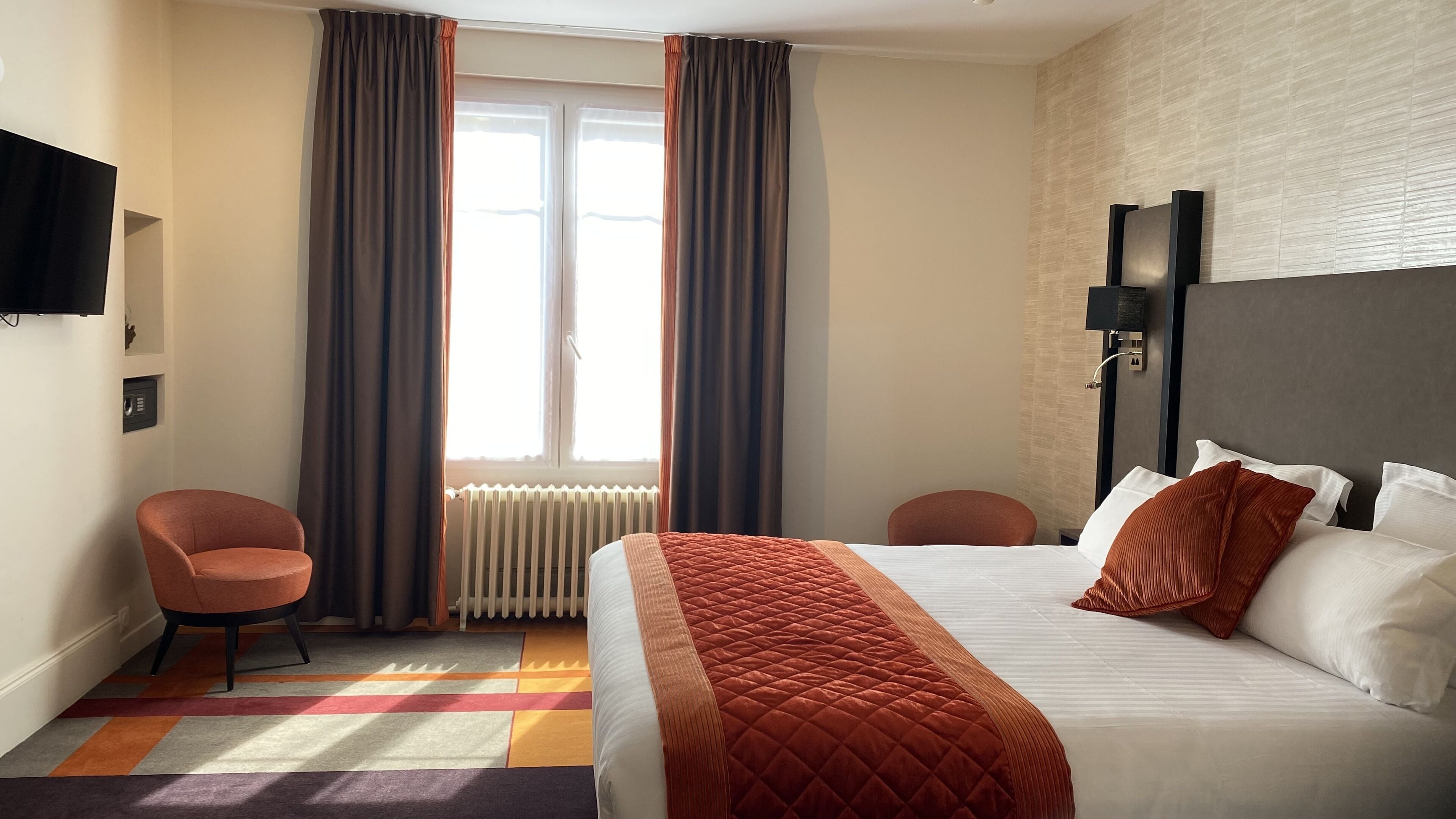 A hotel room with a bed, two chairs, a radiator, curtains, a television, and a wall-mounted light fixture.