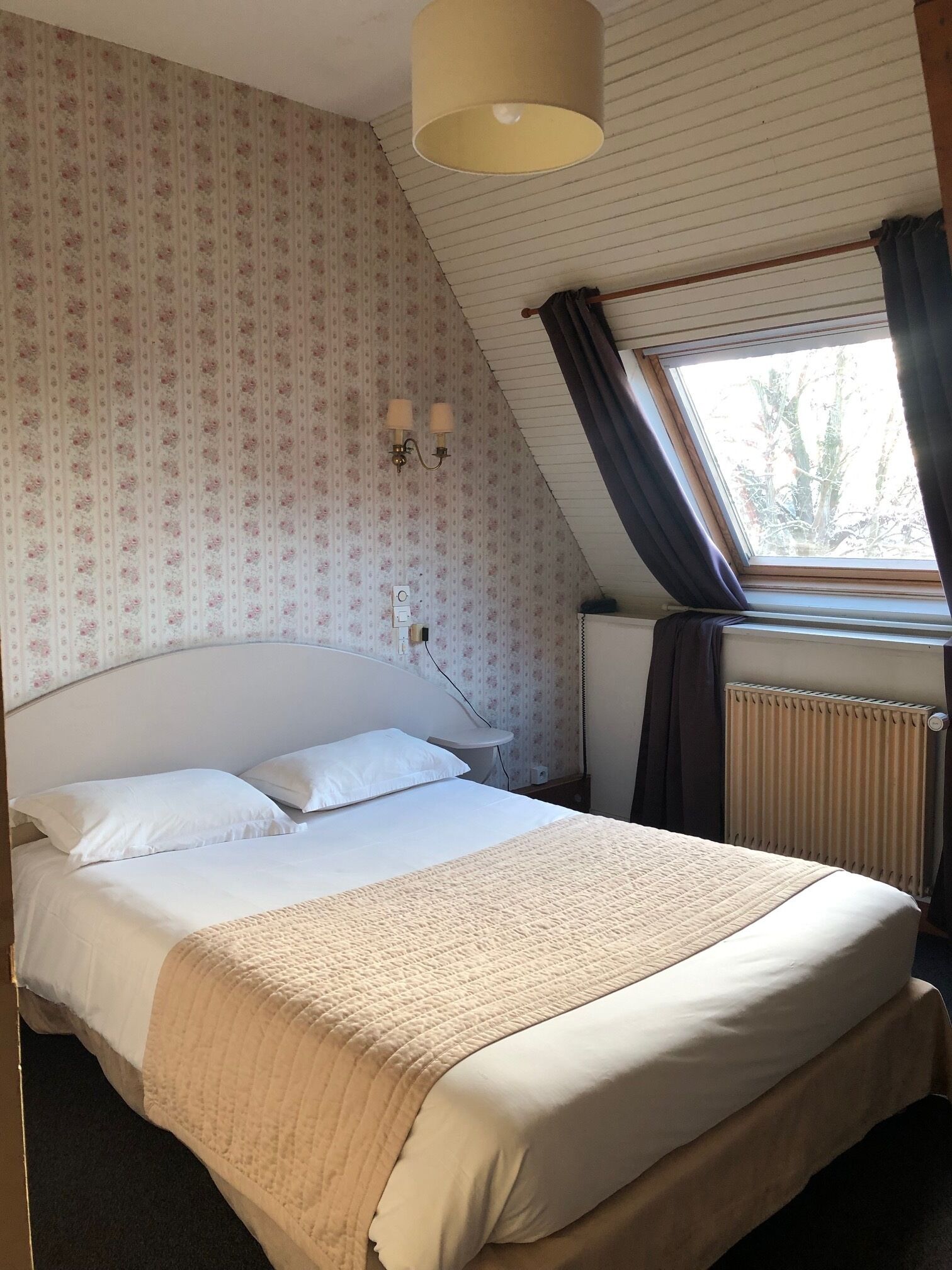 A bedroom with a large bed, a window with curtains, a radiator, and a wall-mounted lamp.