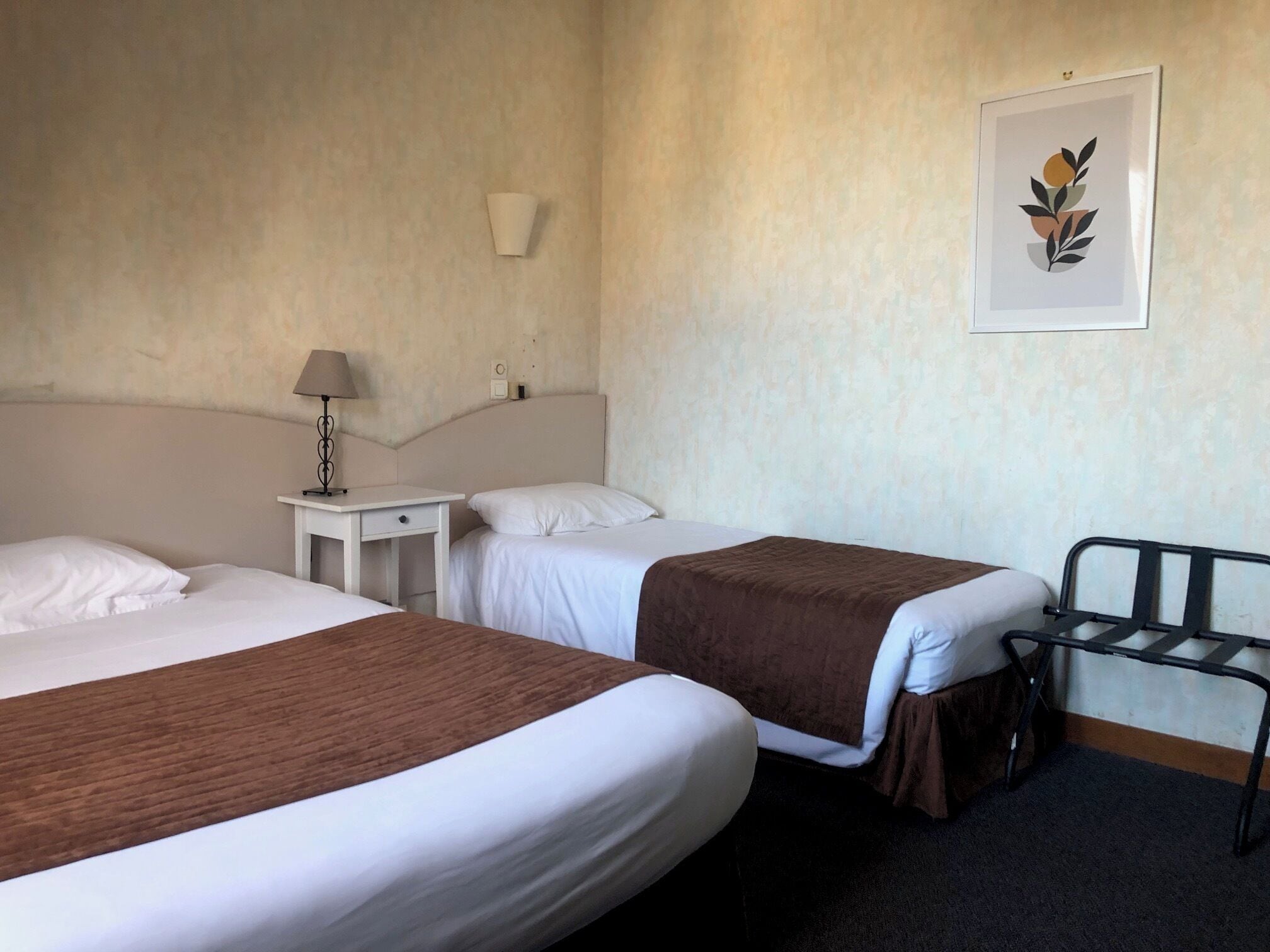 A hotel room with two beds, a desk, a chair, and a wall-mounted lamp.