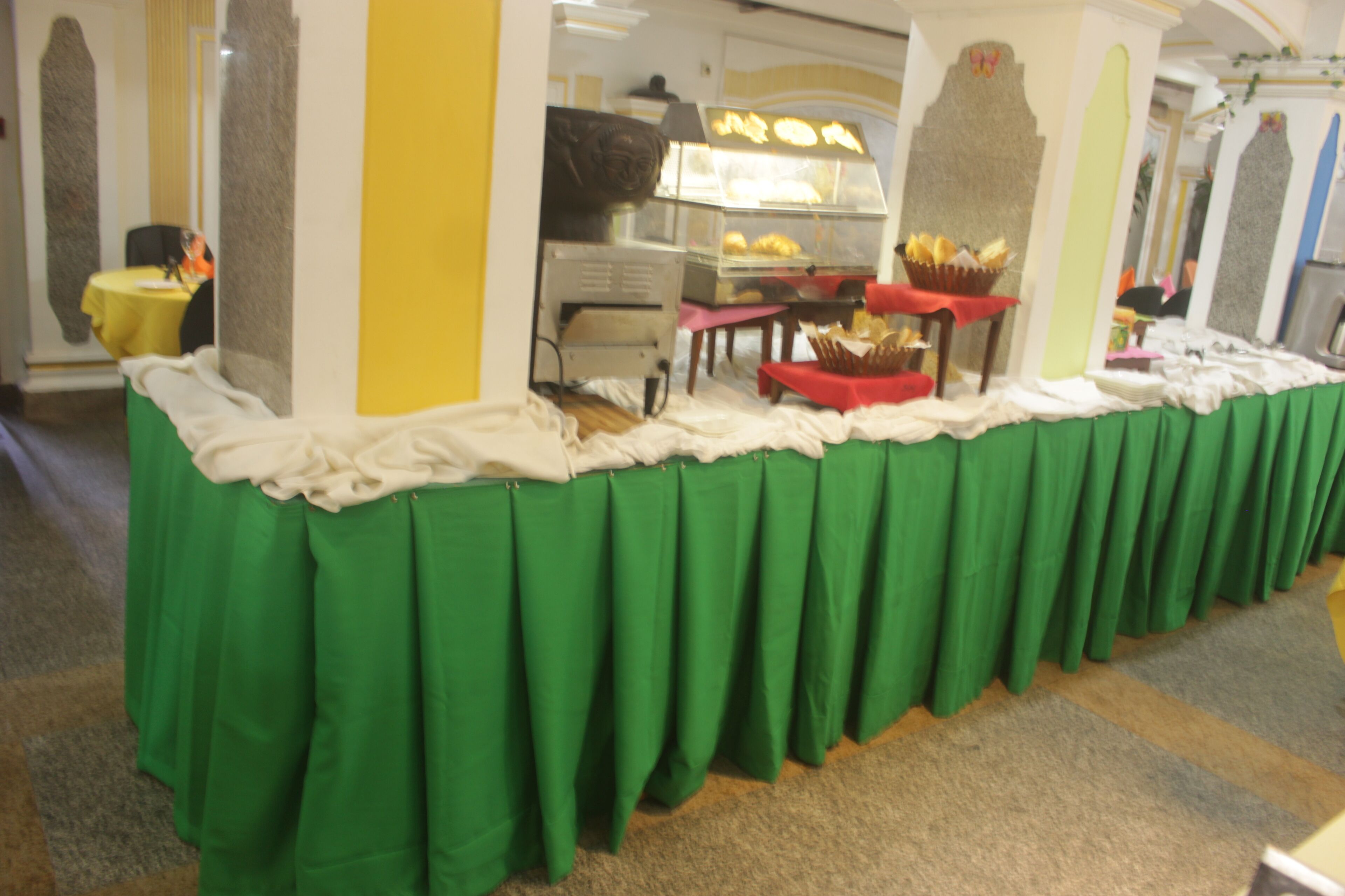 A buffet setup with a green tablecloth, a display case with baked goods, and a red table with napkins.
