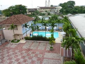 A pool area with palm trees, a building with a tiled roof, and a distant church. - Somatel Hotel (Douala)
