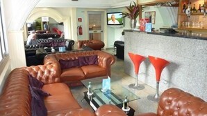 A spacious living area with a large brown leather sofa, a glass coffee table, and a bar area with red bar stools. - Somatel Hotel (Douala)