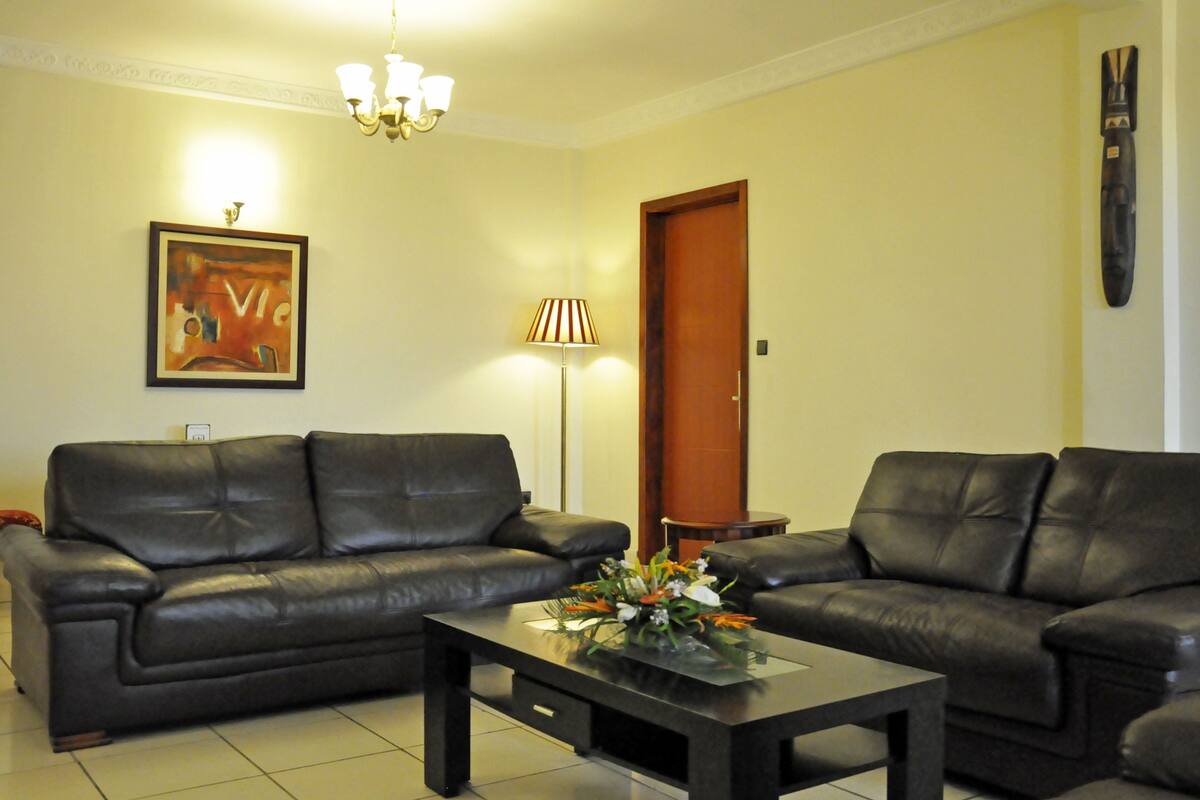 A living room with two black leather sofas, a coffee table with a floral centerpiece, a lamp, and a framed artwork on the wall.