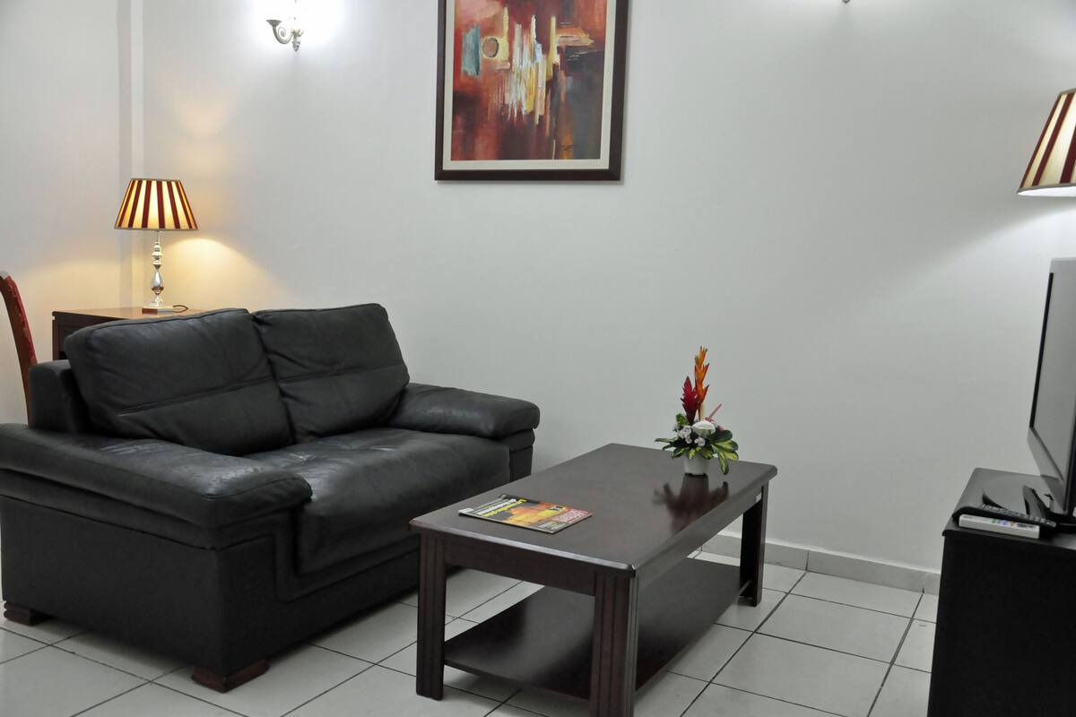 A modern living room with a black leather sofa, a wooden coffee table with a magazine, a lamp, a framed abstract painting, and a TV on a stand.