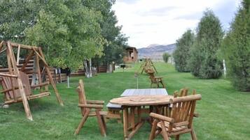 Een houten schommelset, een picknicktafel met stoelen, en een houten speelhuisje in een grasveld met bomen en bergen op de achtergrond.