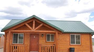 A wooden cabin with a green roof, a front porch with railings, and two windows with shutters.