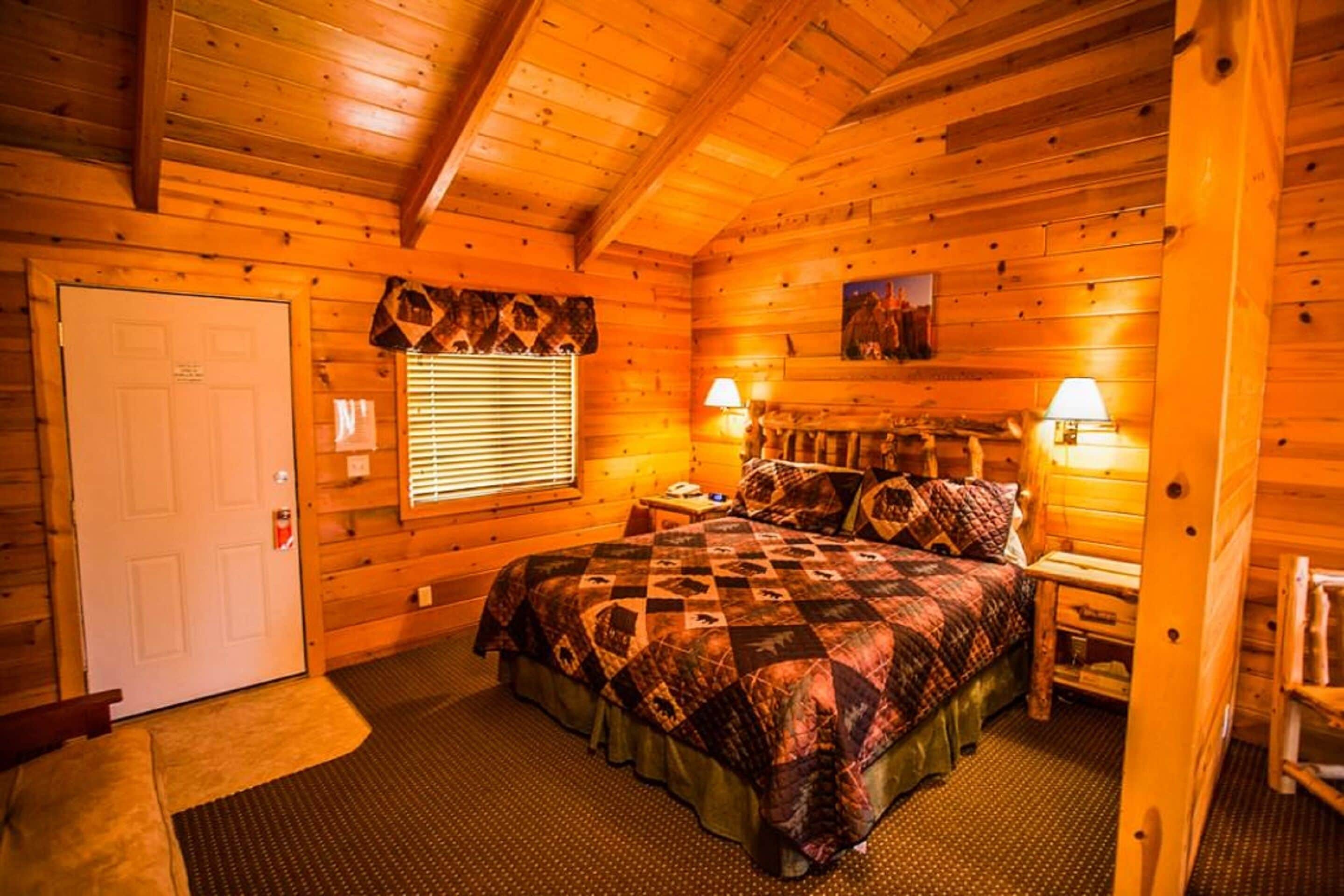 A log cabin bedroom with a bed, two bedside lamps, a window with blinds, and a door.
