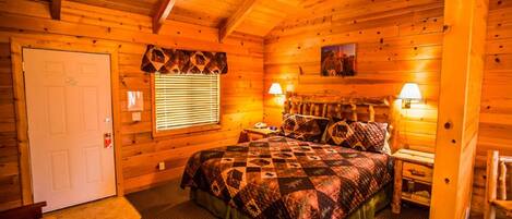 A log cabin bedroom with a bed, two bedside lamps, a window with blinds, and a door.