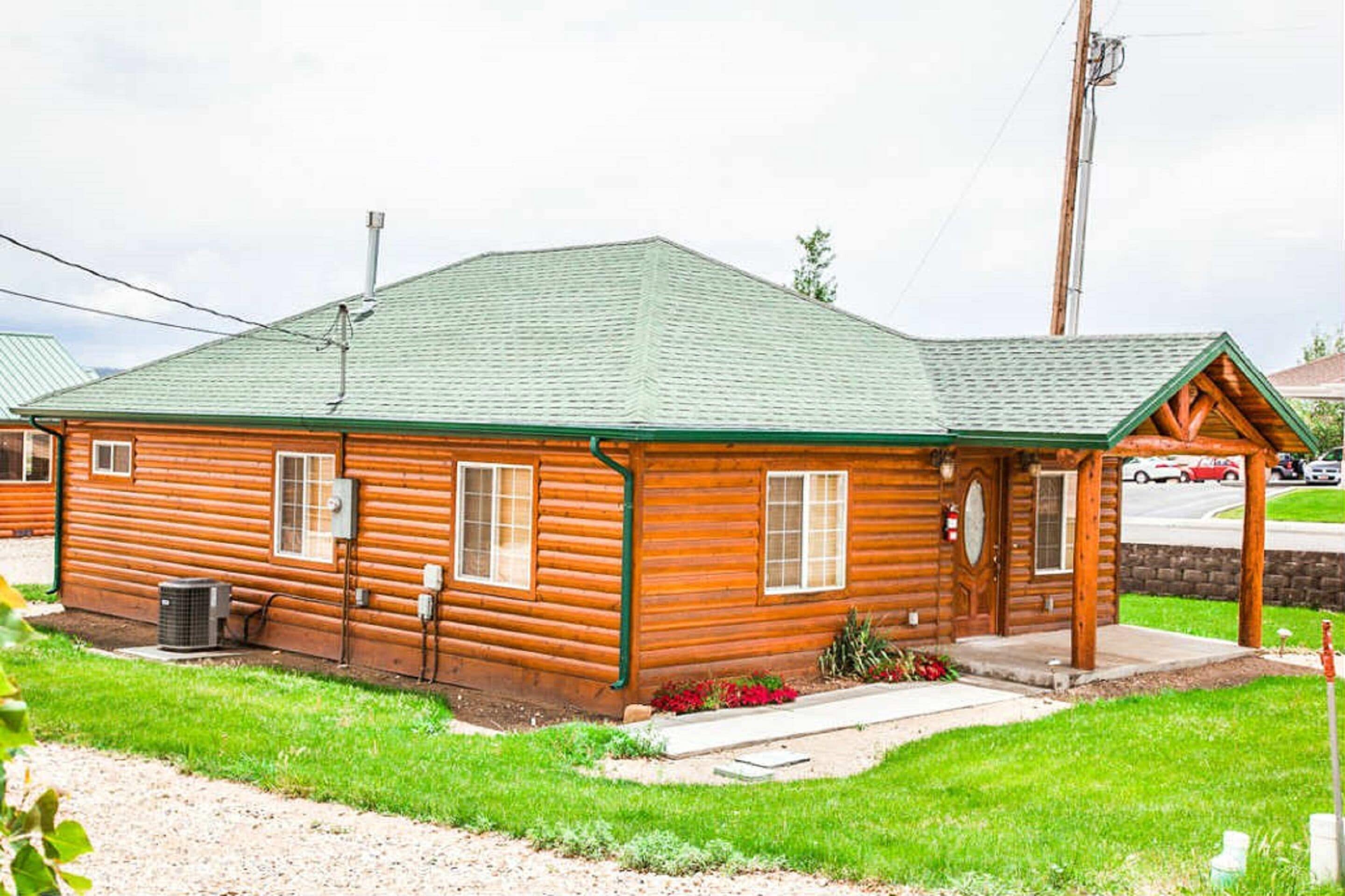 A wooden cabin with a green roof, a front porch, and a small garden area.