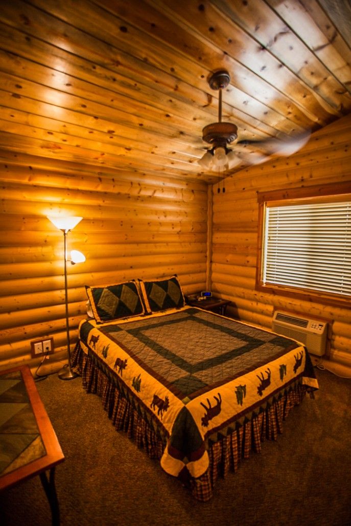 A log cabin room with a bed, a table, a lamp, and a ceiling fan.