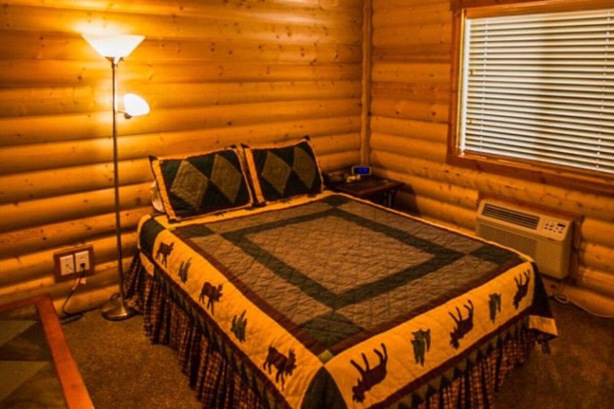 A log cabin bedroom with a bed, a desk, a lamp, and a window.