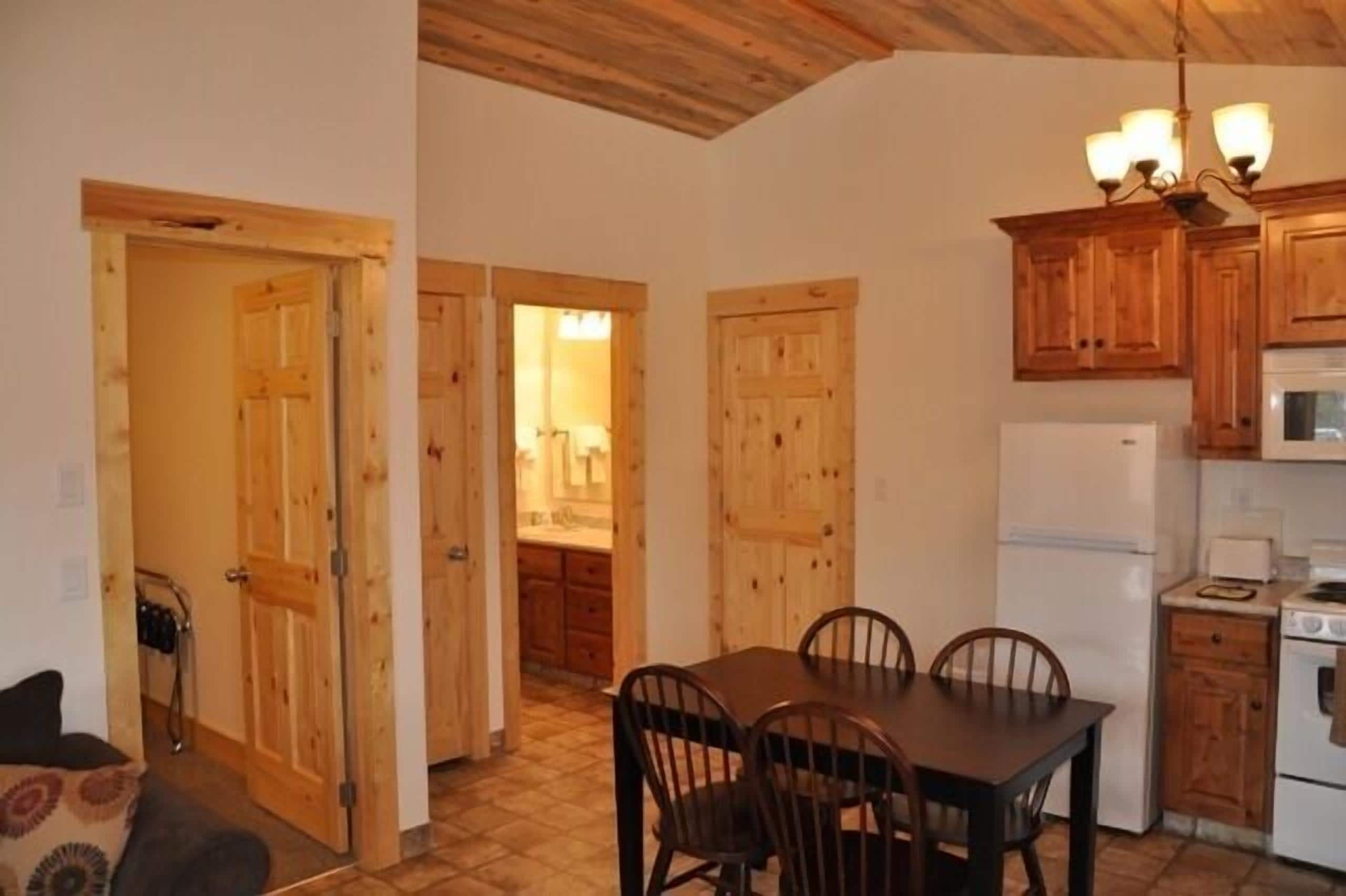 A compact kitchen with wooden cabinets, a dining table with chairs, and a refrigerator.