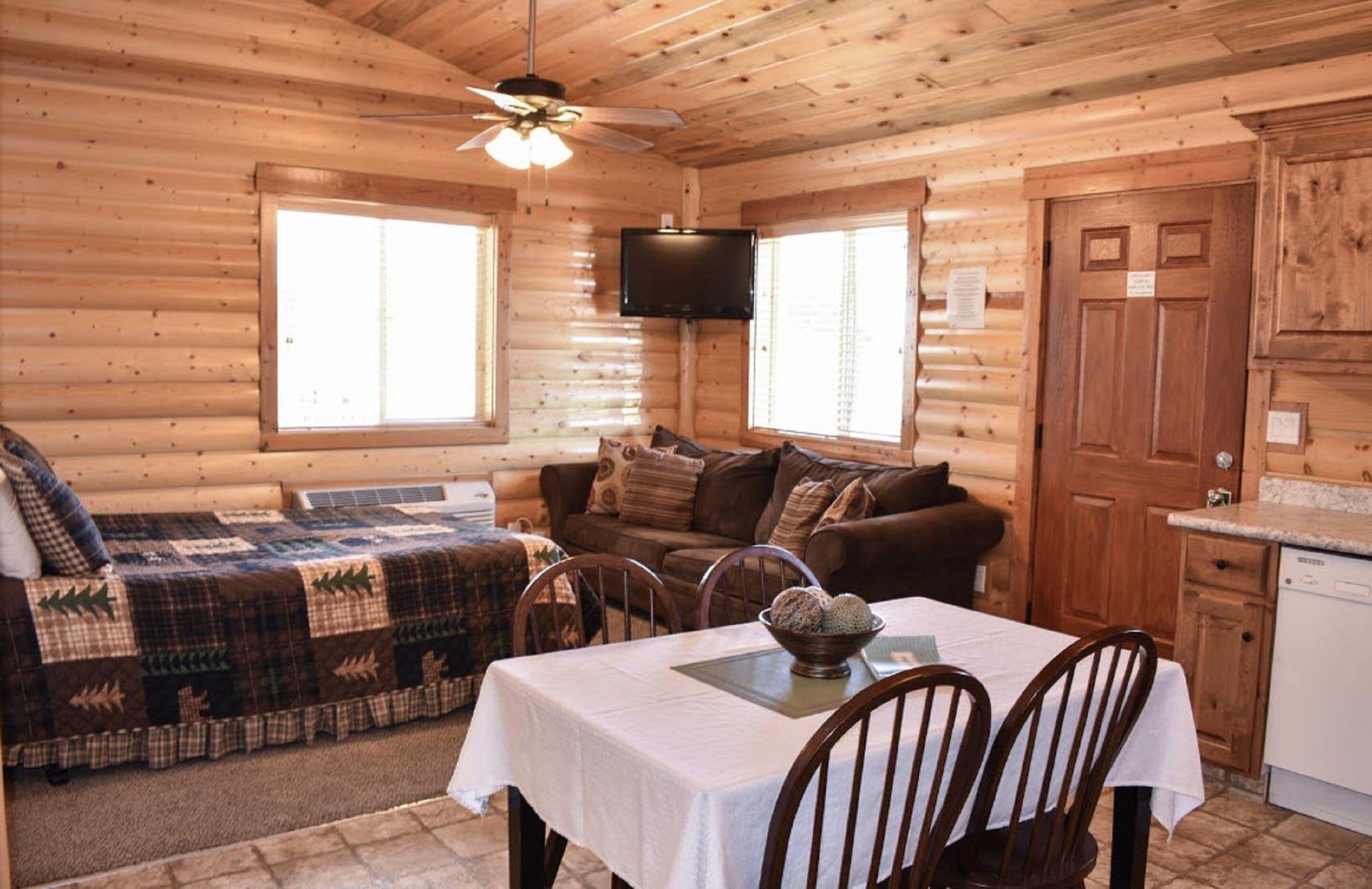 A cozy cabin interior with a dining area, a sofa, and a television mounted on the wall.
