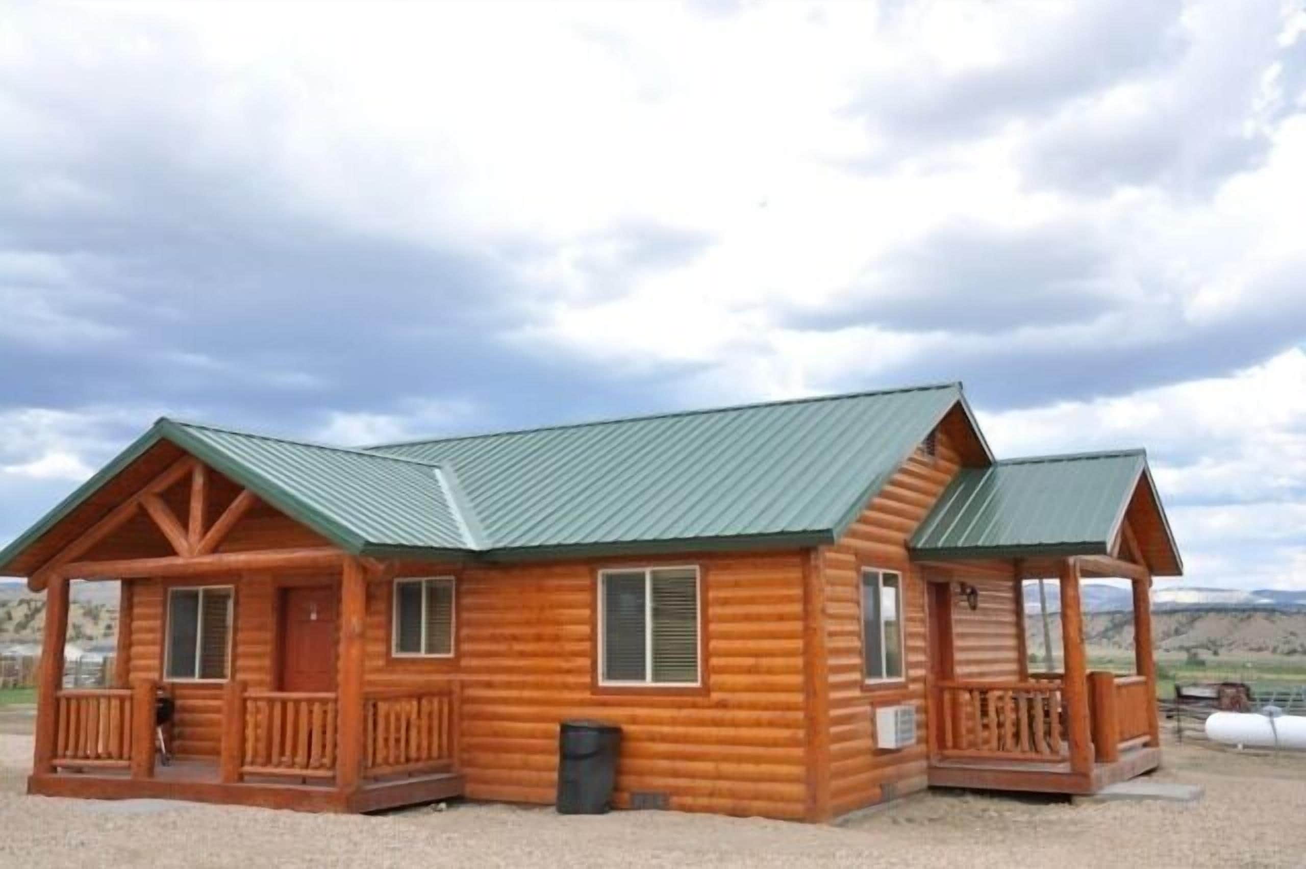 Eine Holzhütte mit grüner Dachdeckung, einer Veranda vorne und einem kleinen Balkon.