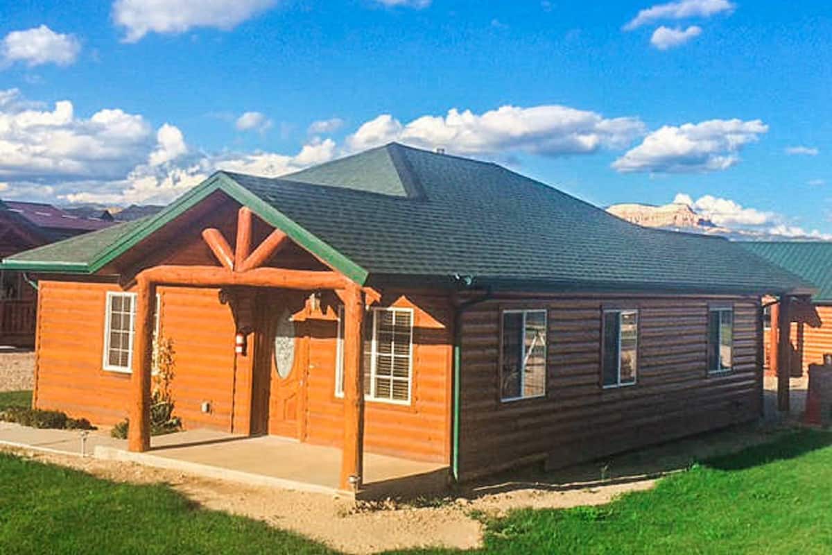 A log cabin with a green roof, a front porch, and a mountain backdrop.