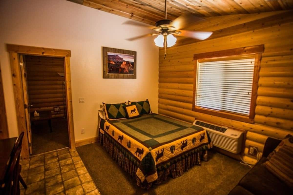 A log cabin bedroom with a bed, a ceiling fan, a window with blinds, and a picture on the wall.