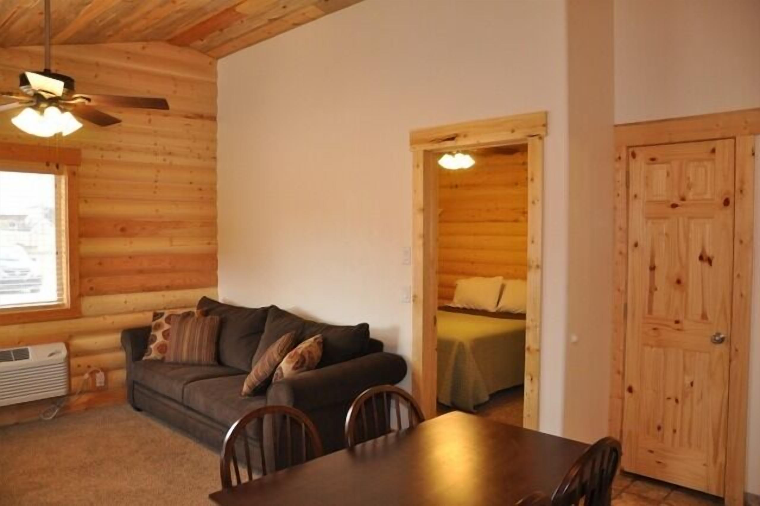 Interior de una cabaña de madera con sofá, mesa de comedor, sillas y cama.