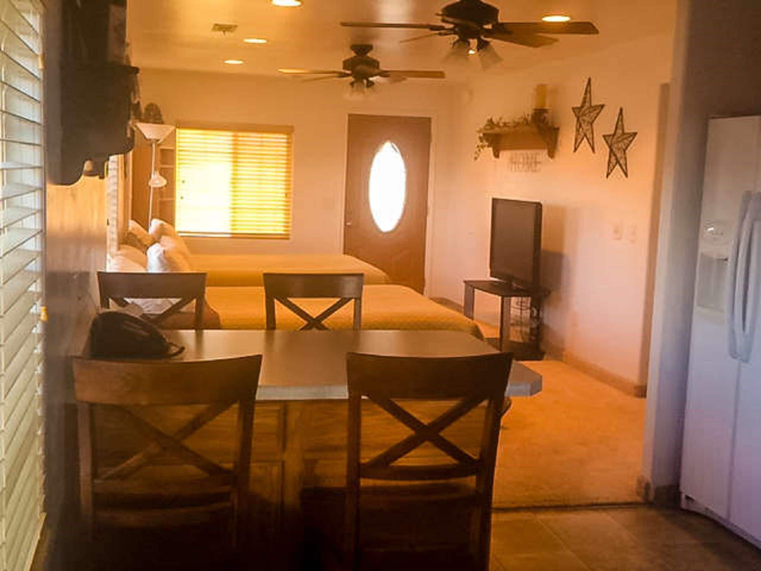 A compact hotel room with a bed, a small dining table with chairs, a ceiling fan, a television, and a refrigerator.
