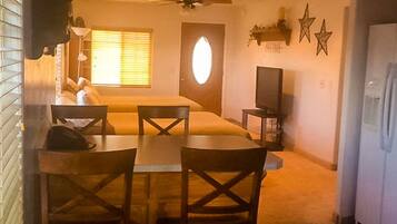 A compact hotel room with a bed, a small dining table with chairs, a ceiling fan, a television, and a refrigerator.