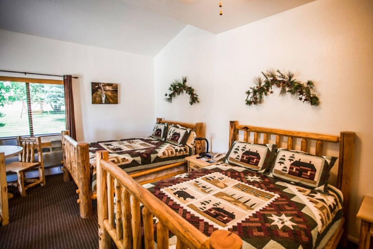 A room with two wooden beds, a small table, and chairs. There is a window with curtains, a picture on the wall, and two wreaths on the wall.