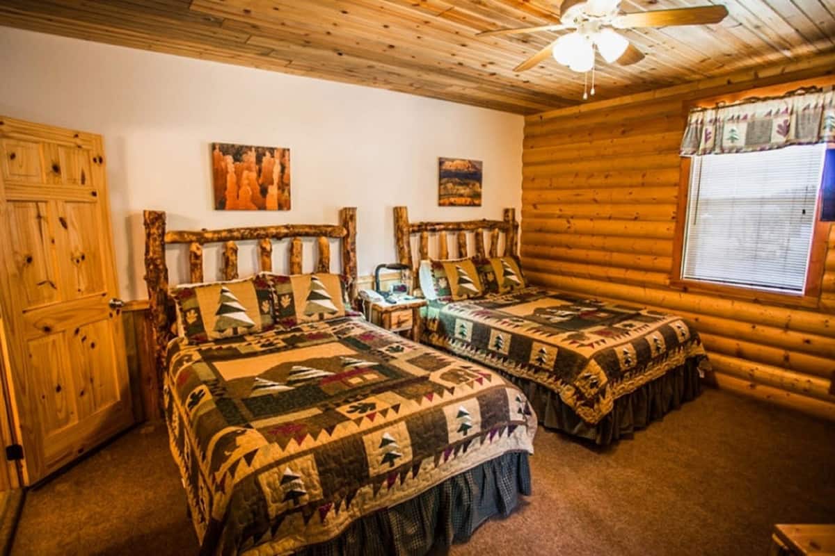 A log cabin bedroom with two beds, wooden walls, a ceiling fan, and a window with curtains.