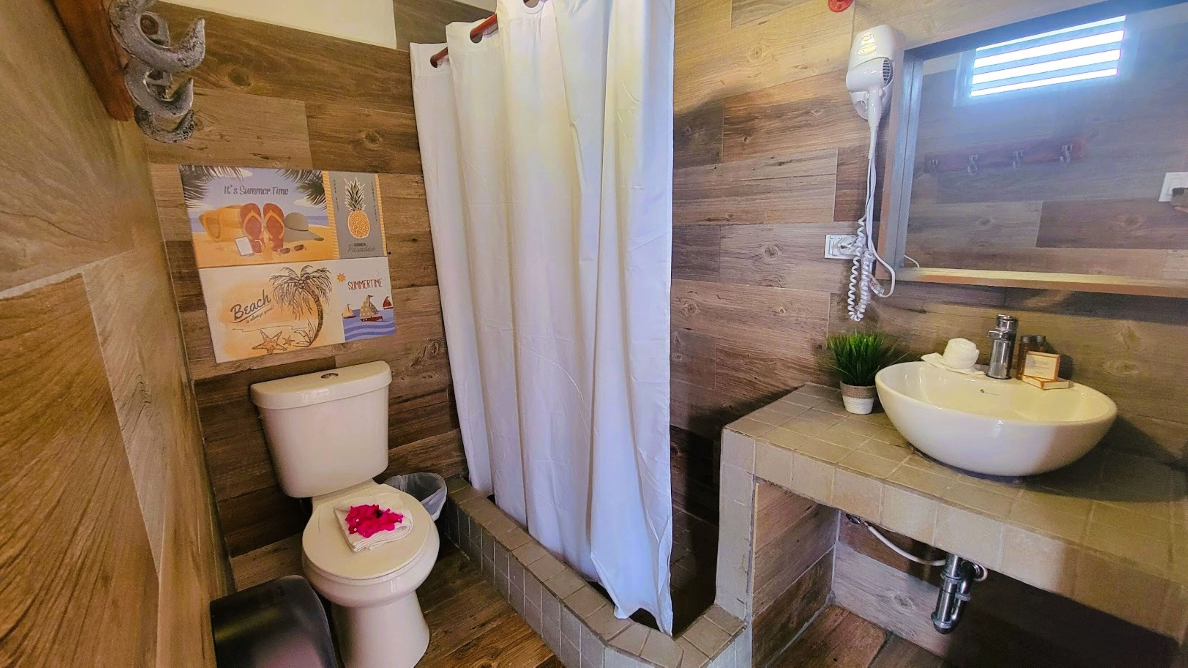 Un baño con suelo de madera, un inodoro blanco, una cortina de ducha, un lavabo con espejo y una pequeña planta en maceta.