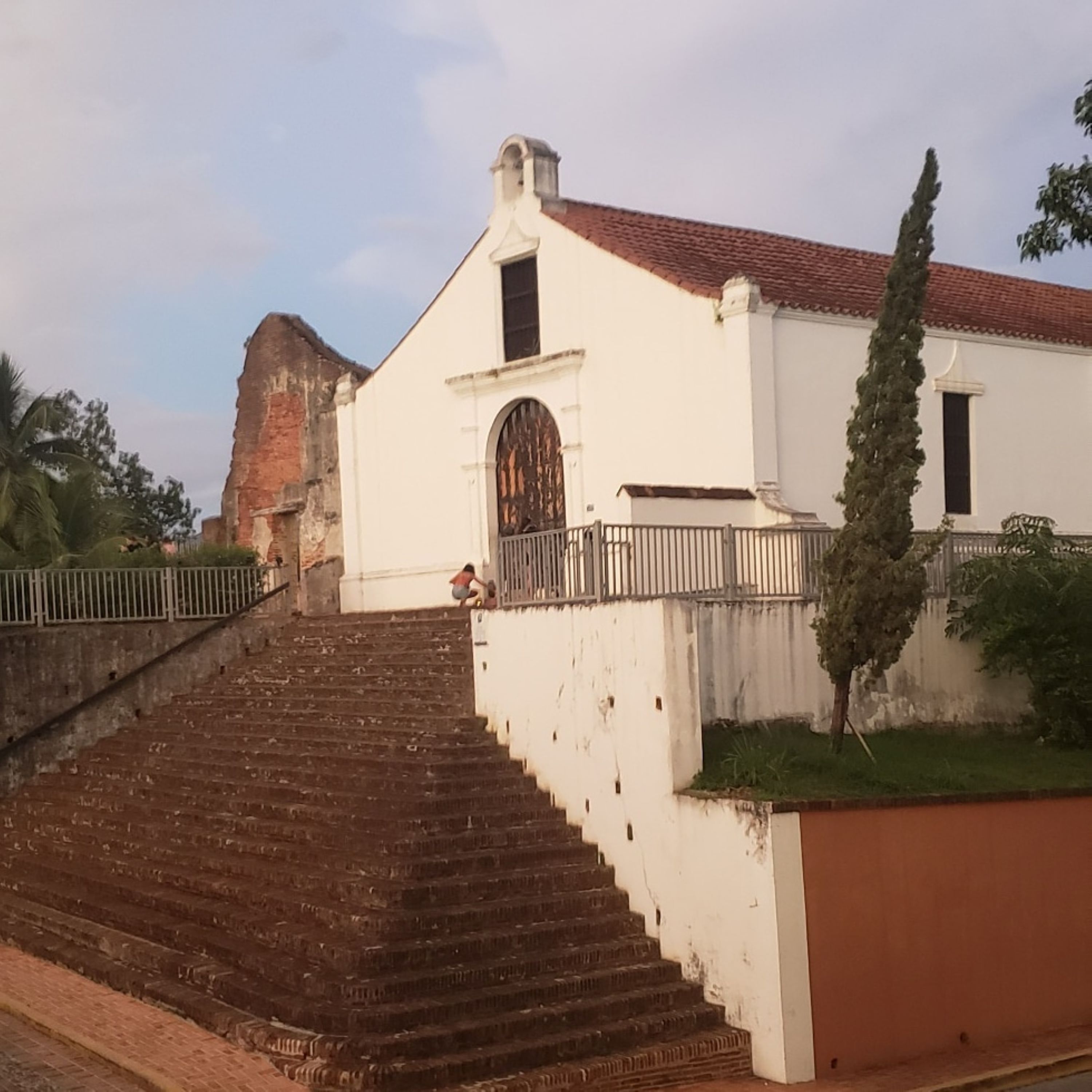 Um edifício com fachada branca e telhado de telhas vermelhas, com uma escadaria que leva a uma entrada com portão.