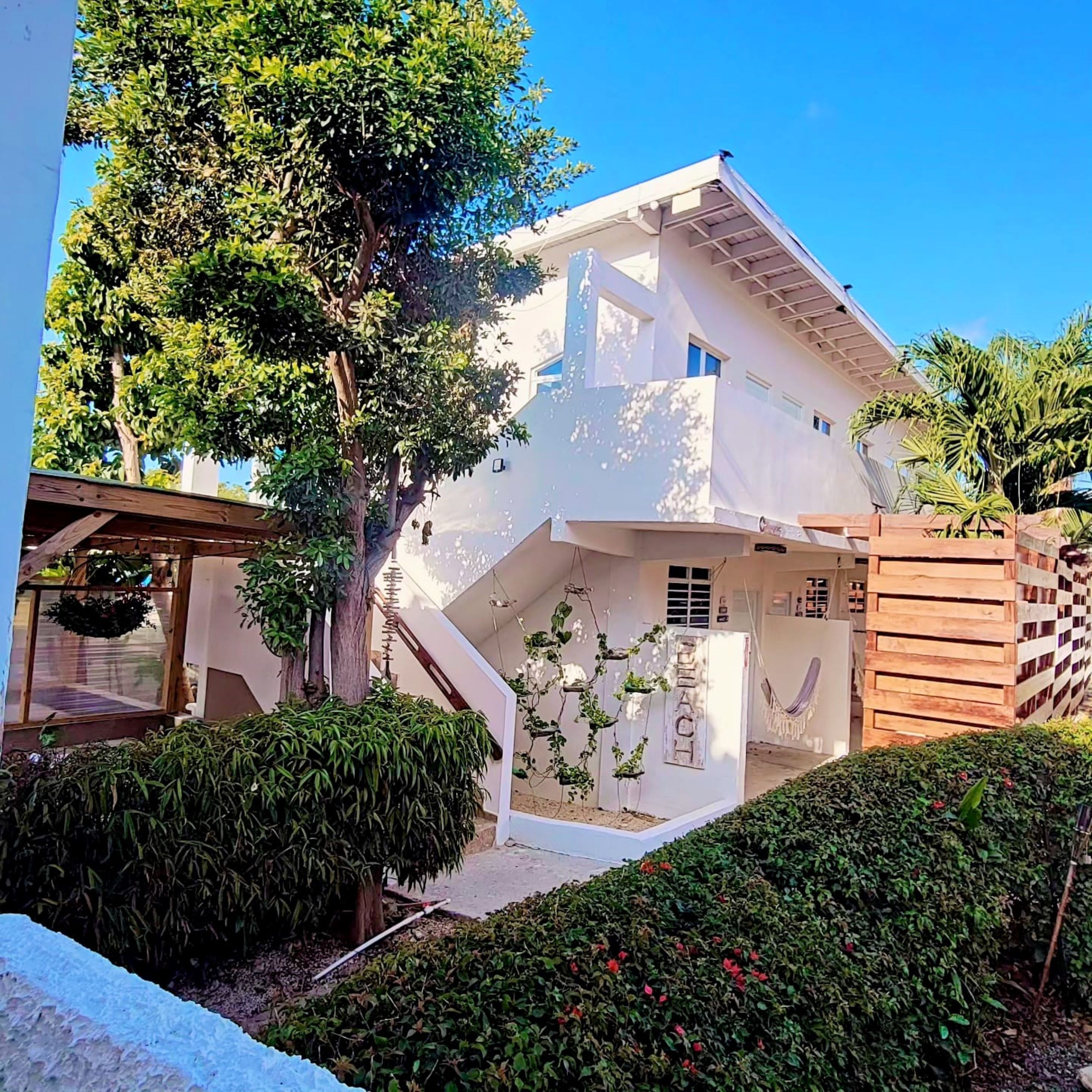 A modern two-story house with a white exterior, a wooden deck, and a small garden area with various plants and a hammock.