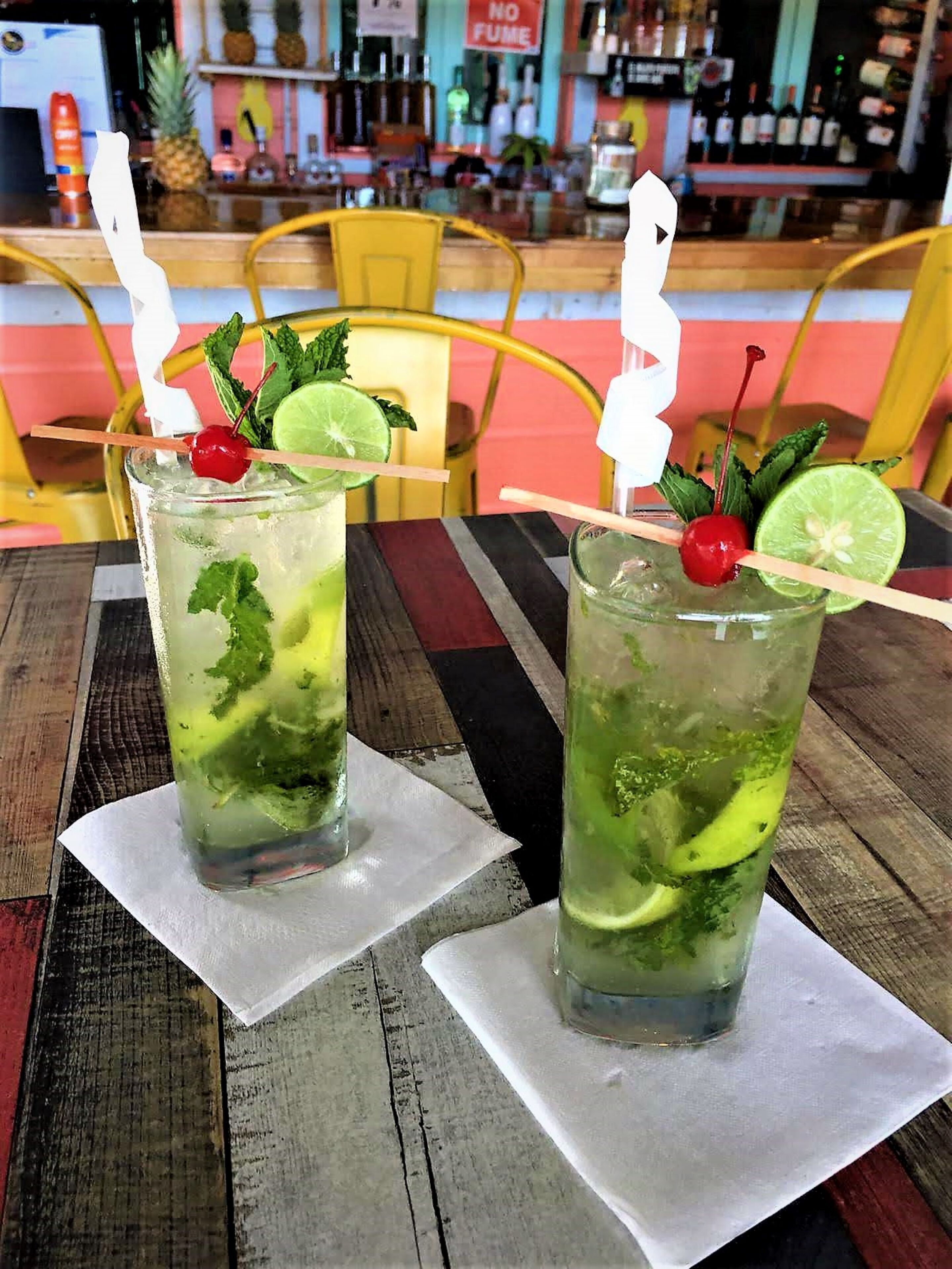 Zwei Gläser mit Limette-Minz-Getränk auf einem Holztisch, im Hintergrund eine Bar.