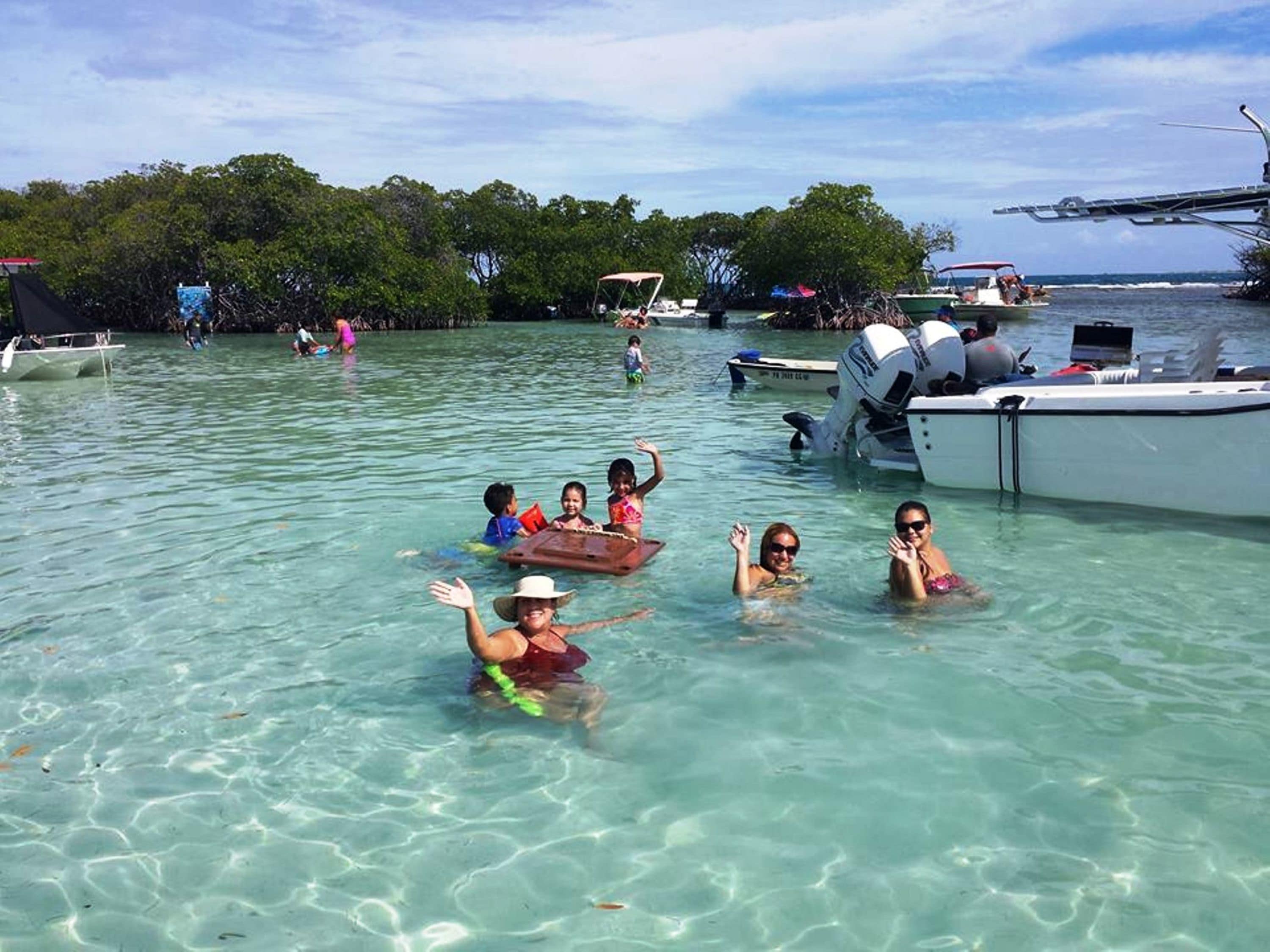 Un grupo de personas en agua cristalina, con barcos y un muelle al fondo.
