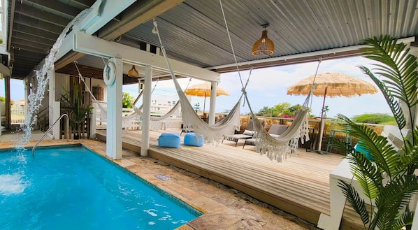 Zona de piscina con una hamaca y sillones de descanso, un porche cubierto y vista al océano.