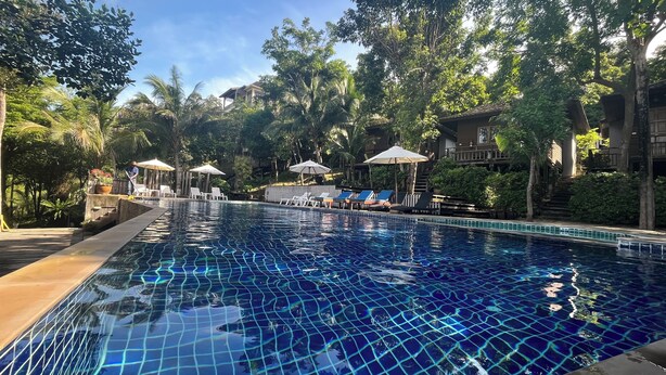 A swimming pool with clear blue water, surrounded by lounge chairs and umbrellas, with trees and buildings in the background. - Chaweng Park Place (Koh Samui)