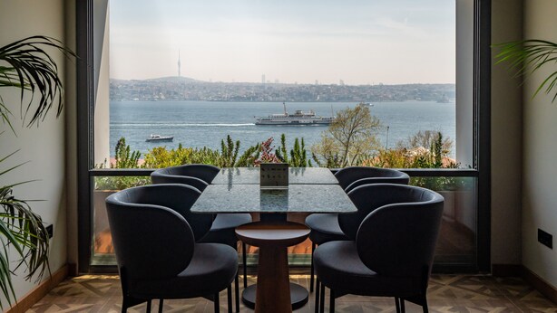 Un comedor con mesa y sillas, vista a un cuerpo de agua y un paisaje urbano al fondo.