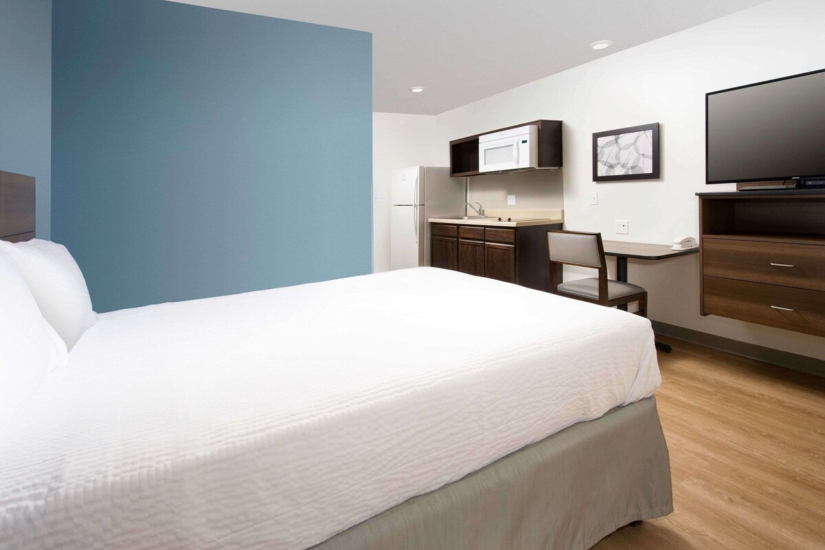 A hotel room with a bed, a desk with a chair, a microwave, a refrigerator, and a flat-screen TV.